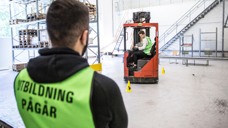 Man i väst med texten Utbildning pågår tittar på man som kör truck