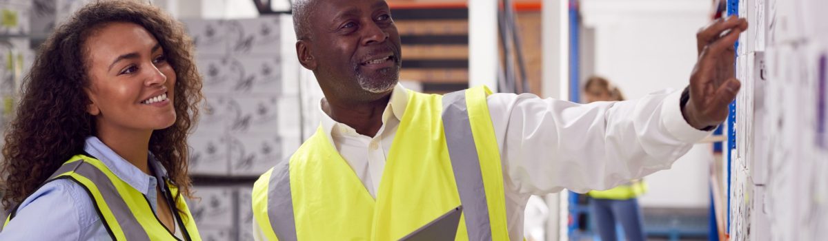 Male Team Leader With Digital Tablet In Warehouse Training Intern Standing By Shelves
