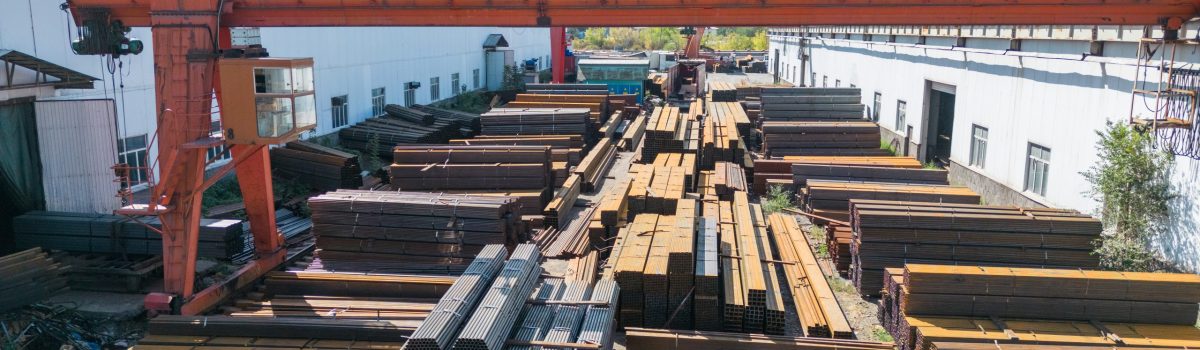 Galvanized iron pipes stack and stainless steel rectangular bars of metal with gantry crane in the background